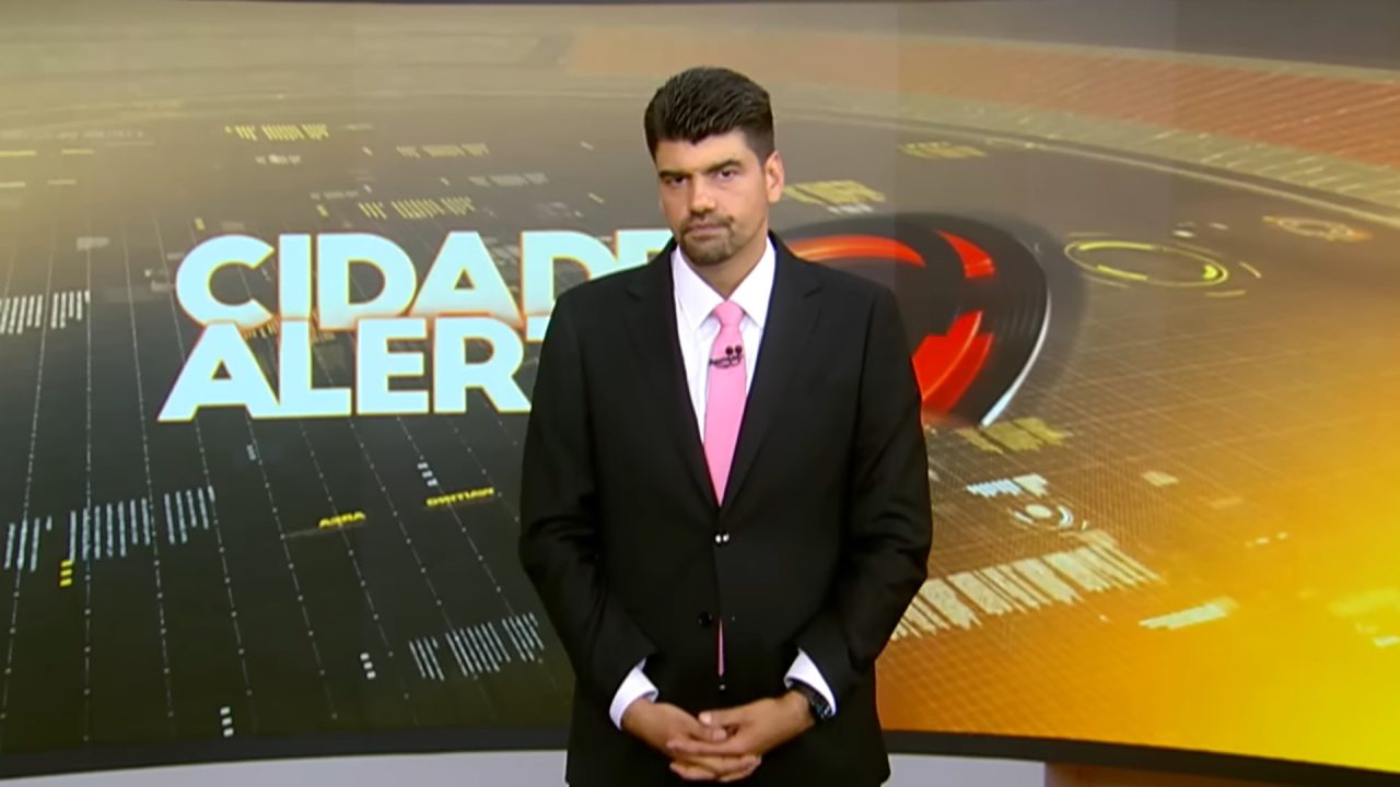 André Azeredo, apresentador do Cidade Alerta, posa de terno e gravata posa diante de telão com logo do programa da Record.