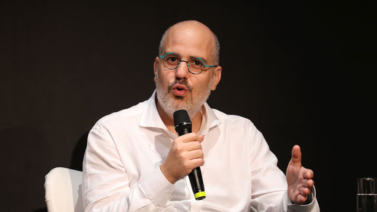 André Basbaum, presidente da EBC, fala ao microfone em evento, homem calvo com barba grisalha, usa óculos redondos e camisa social branca, fundo preto