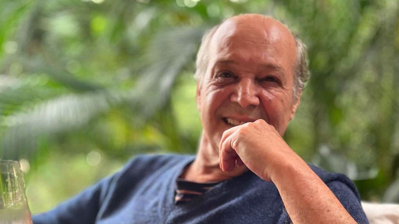 André de Biase sorri enquanto posa sentado em ambiente externo com vegetação ao fundo