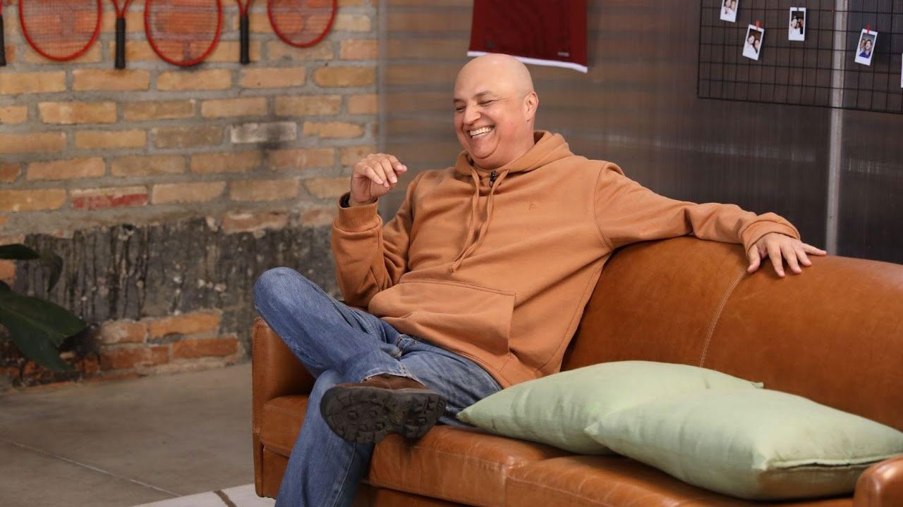 André Henning sorrindo sentado em sofá durante gravação em ambiente descontraído com decoração esportiva