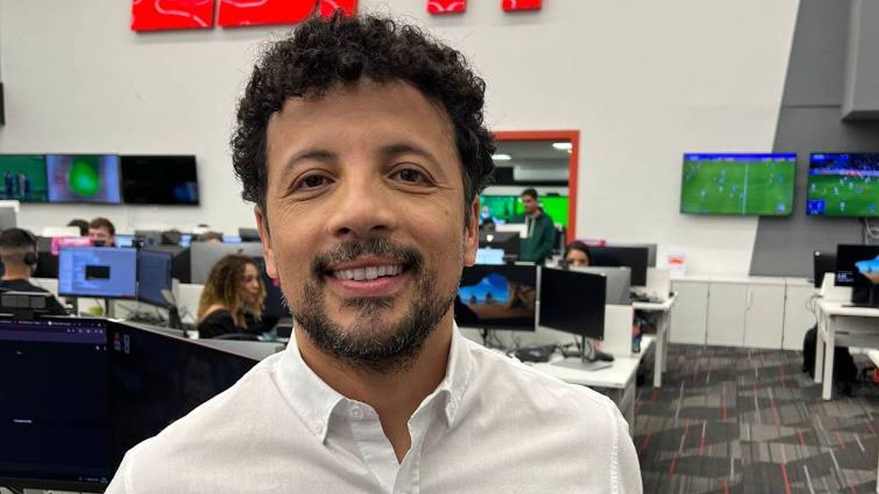 André Hernan sorri de camisa branca na redação da ESPN com computadores e telas exibindo partidas de futebol ao fundo