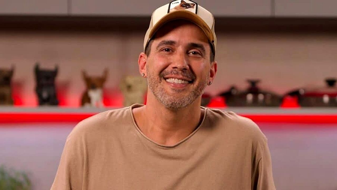 Um homem aparece em um ambiente interno, sorrindo diretamente para a câmera. Ele usa boné claro, camiseta bege e tem barba curta grisalha. Ao fundo, há uma prateleira com objetos decorativos e iluminação em tom avermelhado, criando um ambiente moderno e descontraído. A composição sugere um cenário de estúdio ou gravação de conteúdo.