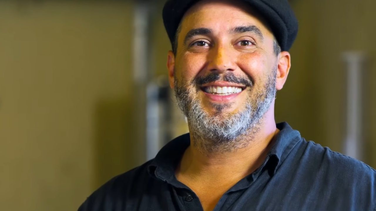 Na imagem, o apresentador André Marques aparece sorridente, usando uma camisa preta e um boné combinando. Ele está em um ambiente interno com iluminação suave, que destaca seu rosto iluminado e expressão descontraída.