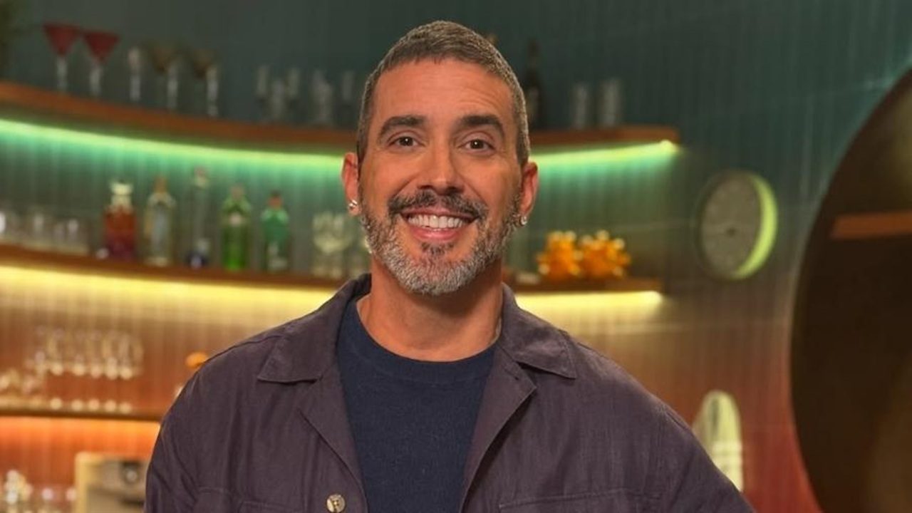 Homem sorridente, com barba curta grisalha e cabelo também grisalho, aparece em ambiente interno iluminado. Ele usa camiseta escura e camisa casual aberta. Ao fundo, há prateleiras com garrafas e copos, além de iluminação colorida que cria clima de estúdio ou bar moderno.