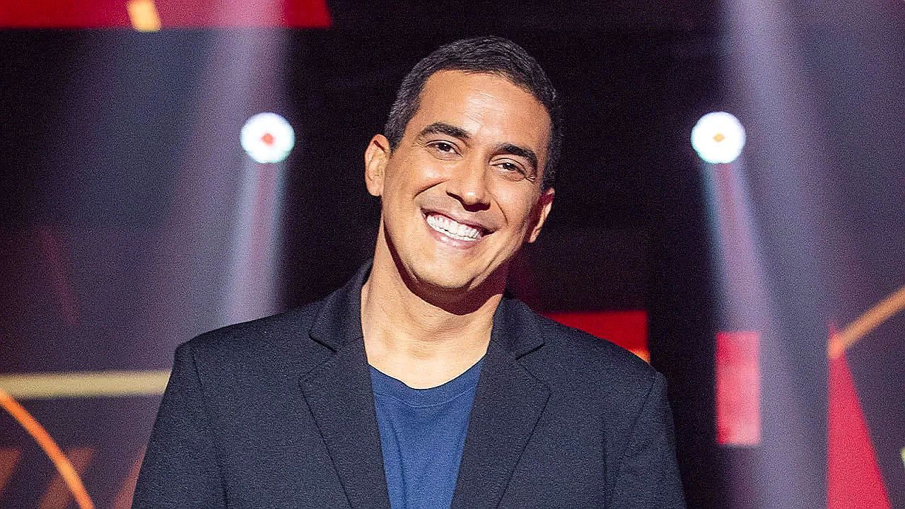 Homem sorri para a câmera em estúdio de televisão, usando blazer escuro sobre camiseta azul. Ao fundo, luzes de palco e elementos gráficos iluminados sugerem cenário de programa de entretenimento ou competição.