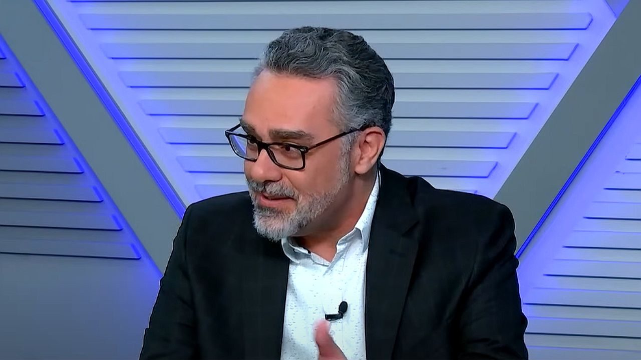 André Marsiglia fala durante programa de debates usando terno preto e óculos em estúdio de televisão