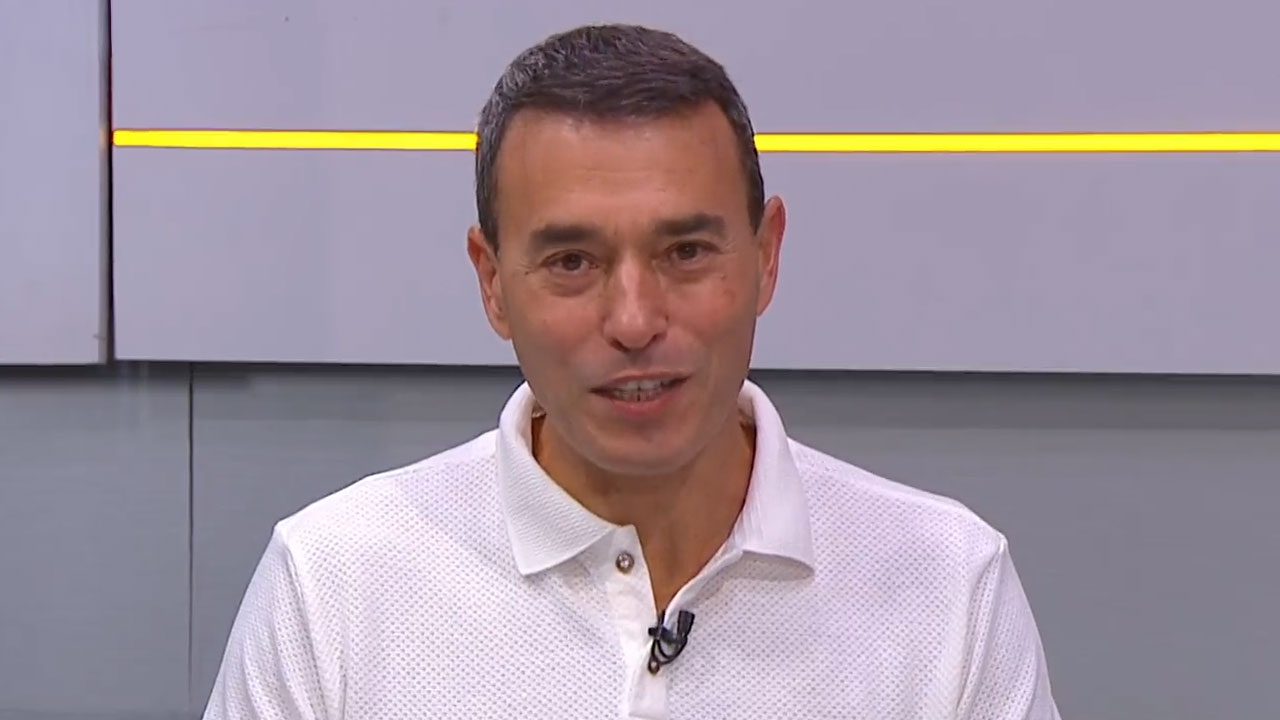O apresentador André Rizek, com camisa polo branca, comanda programa de esporte no estúdio do SporTV com painel cinza e faixa amarela