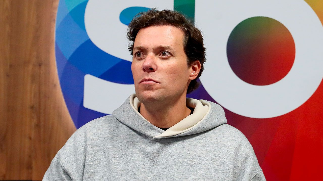 André Valadão de moletom cinza e capuz, em plano médio, olhando para o lado no fundo com o logo do SBT