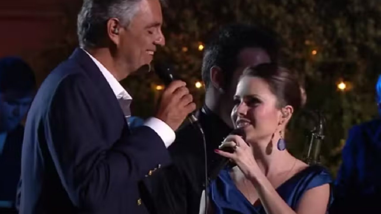 A imagem mostra um homem e uma mulher cantando juntos em um palco, cada um segurando um microfone. Eles parecem estar sorrindo um para o outro, transmitindo sintonia e emoção durante a apresentação. Ao fundo, há músicos e iluminação suave, sugerindo um show ao vivo em ambiente elegante