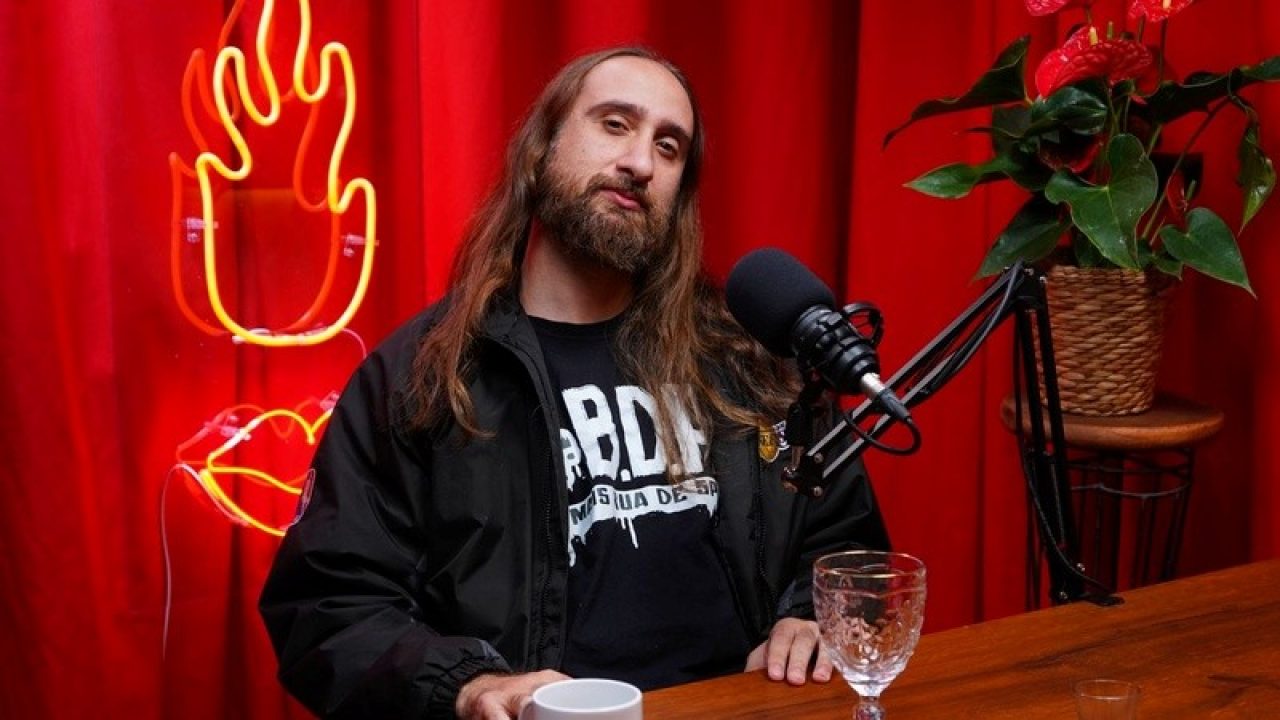 Andreas Ciero de cabelos longos e barba em estúdio de podcast com fundo de cortina vermelha, vestindo jaqueta preta sobre camiseta de estampa branca