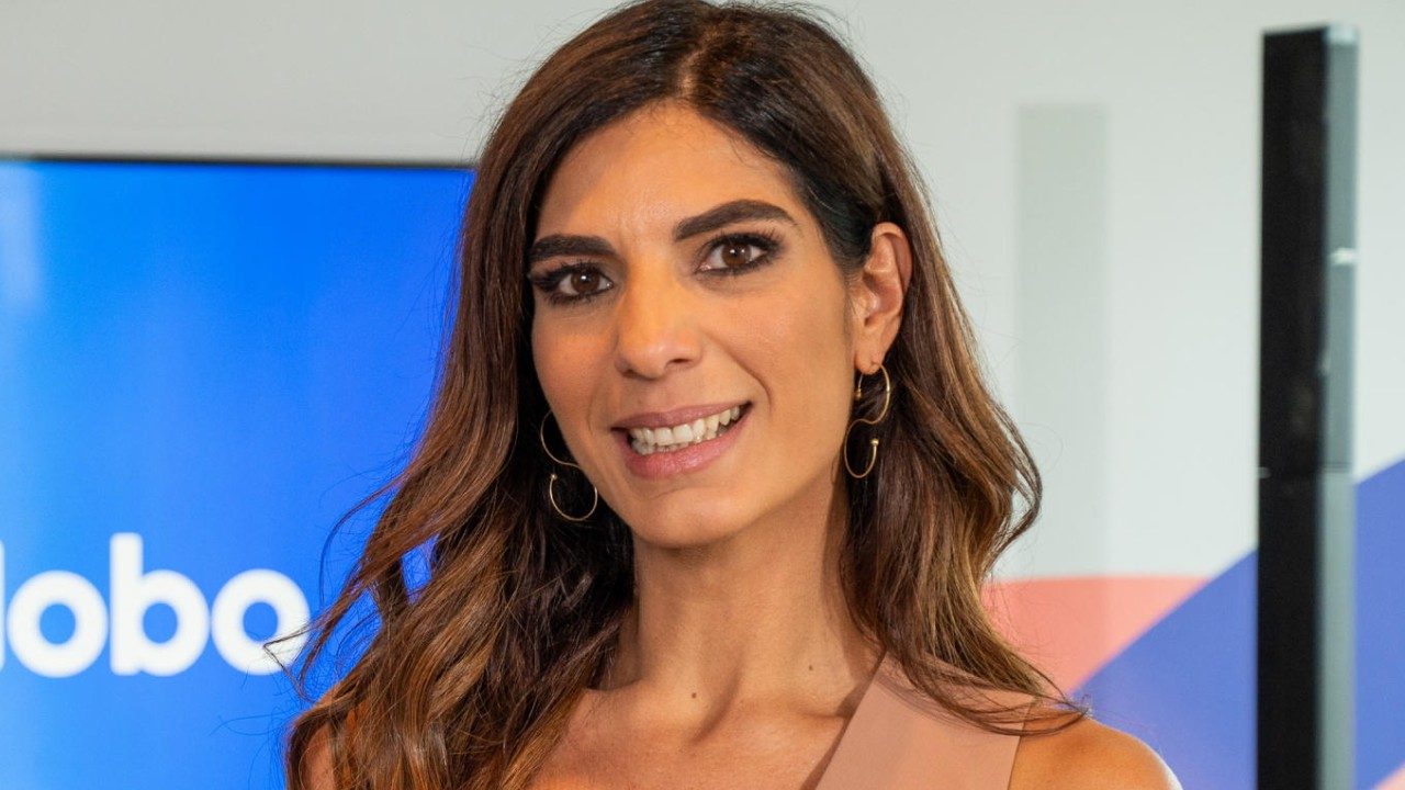 Andréia Sadi sorri em estúdio de TV com monitor com logo da Globo ao fundo e brincos de argola visíveis