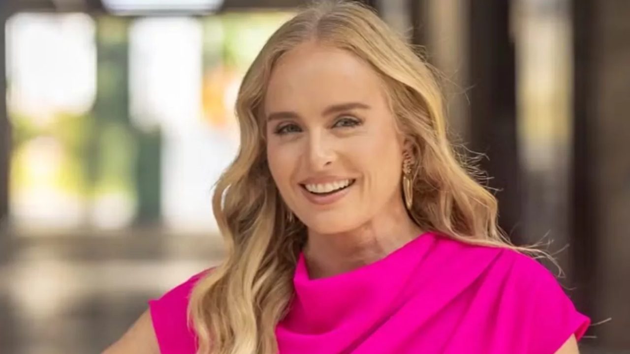 Angélica de cabelos ondulados sorri usando vestido rosa vibrante e brincos dourados em ambiente iluminado com fundo desfocado