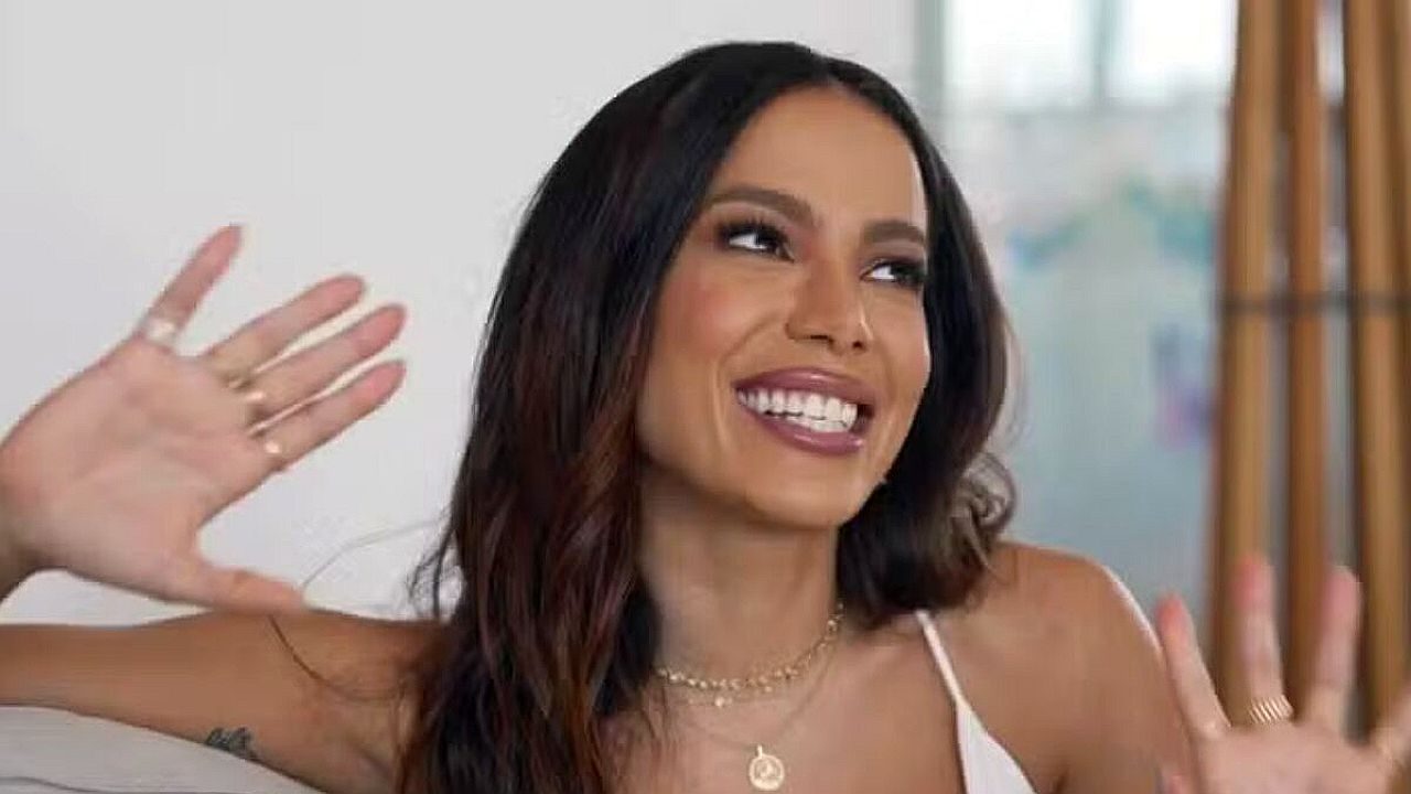 Mulher de cabelo longo e escuro sorri amplamente enquanto gesticula com as mãos erguidas, em conversa descontraída. Ela usa blusa clara e colares delicados, sentada em ambiente interno bem iluminado, com expressão animada e espontânea.