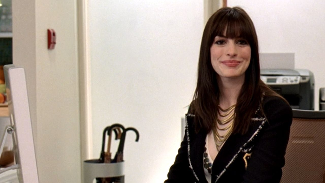 Mulher de cabelo castanho com franja sorri sentada em escritório com computador e guarda-chuvas ao fundo