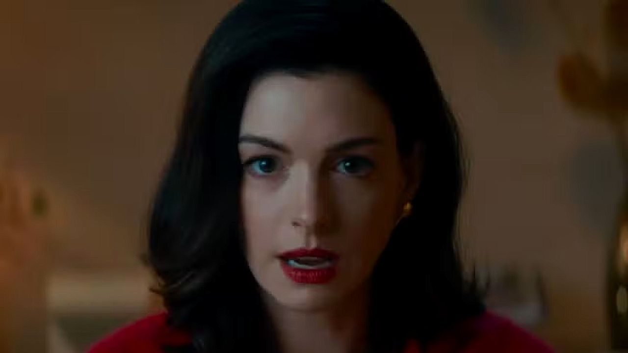 Anne Hathaway com expressão séria e batom vermelho em cena de filme durante conversa em ambiente interno