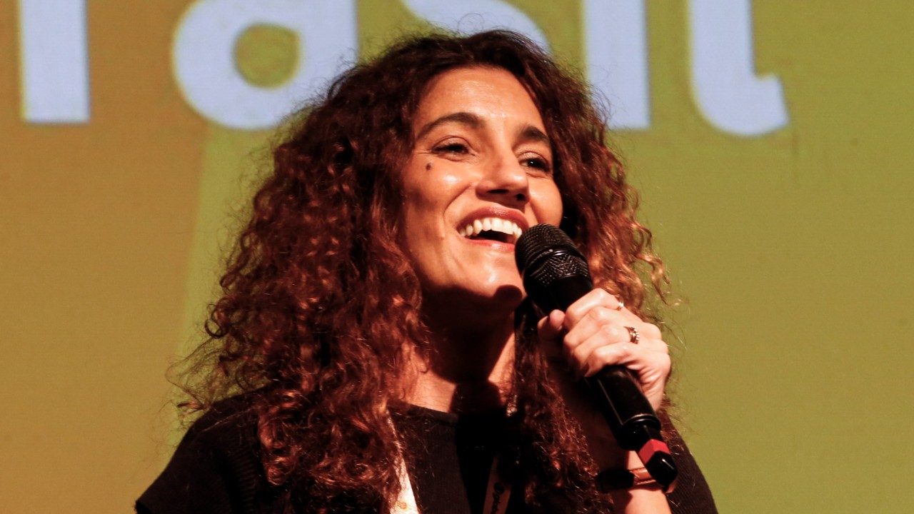 Antonia Pellegrino sorri enquanto fala ao microfone em palco com fundo iluminado durante evento