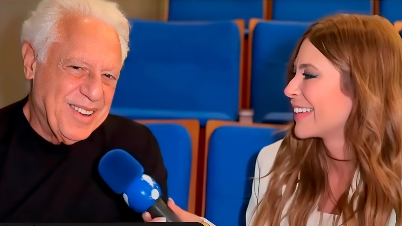 Antonio Fagundes fala ao microfone azul enquanto Naty Curvelo sorri durante entrevista em auditório com poltronas azuis ao fundo