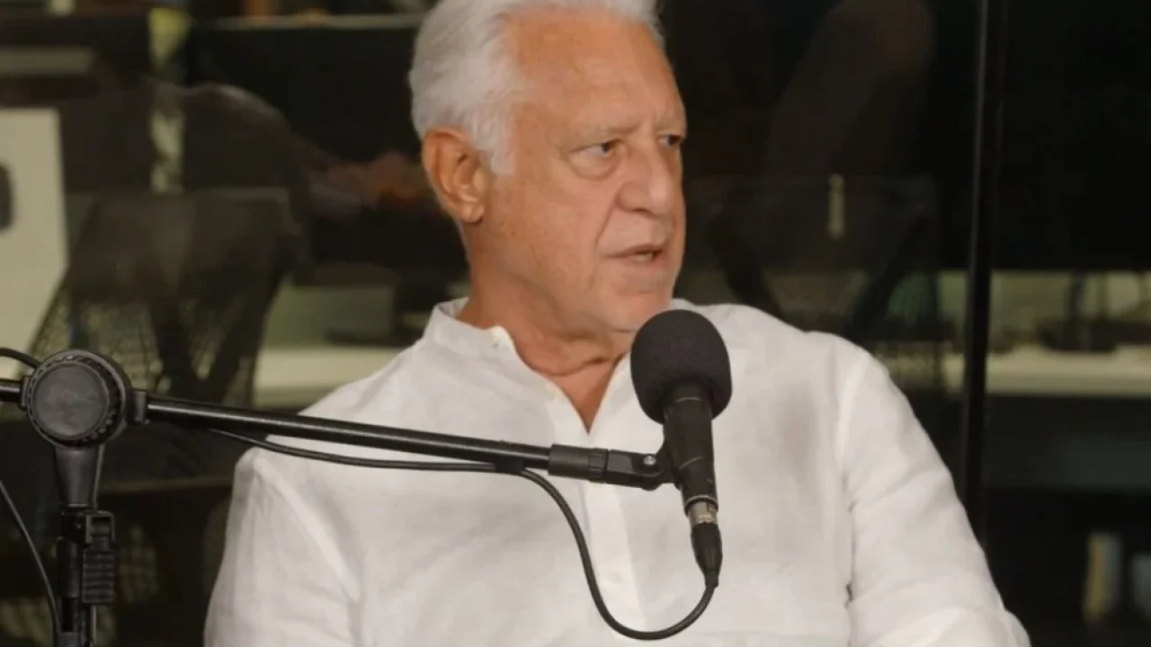A imagem mostra o ator Antonio Fagundes durante uma entrevista em um videocast, sentado em frente a um microfone profissional. Vestindo uma camisa branca de linho, ele aparece com uma expressão séria e atenta, demonstrando envolvimento na conversa.