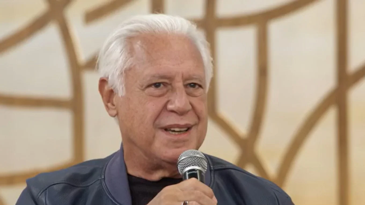 Homem mais velho, de cabelos brancos e expressão serena, segura um microfone enquanto fala em um ambiente de estúdio iluminado com fundo abstrato em tons de bege e dourado. Ele veste jaqueta azul e camisa escura, transmitindo postura confiante e acolhedora típica de entrevistas ou participações em programas de TV.