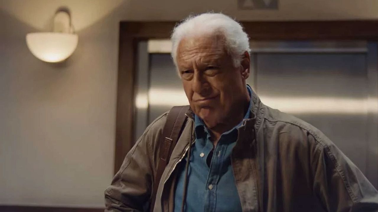 Homem idoso de cabelos brancos com mochila observa com expressão séria em corredor interno próximo a elevador