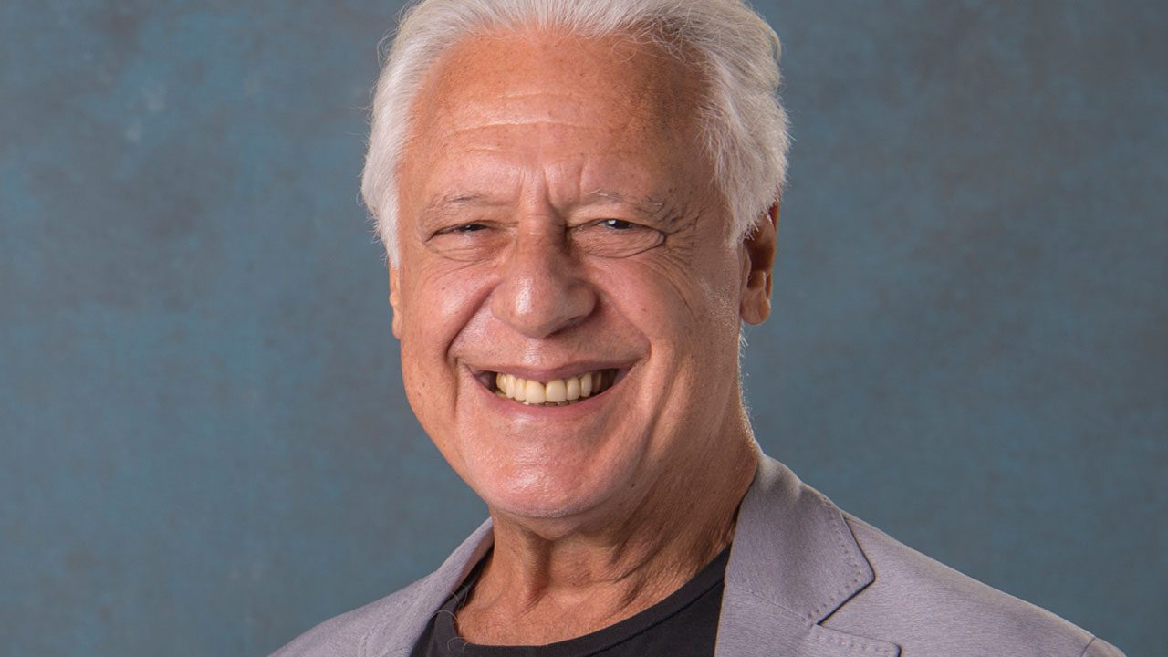 Ator Antônio Fagundes sorrindo, com cabelo branco e casaco cinza claro, em foto de divulgação da Globo