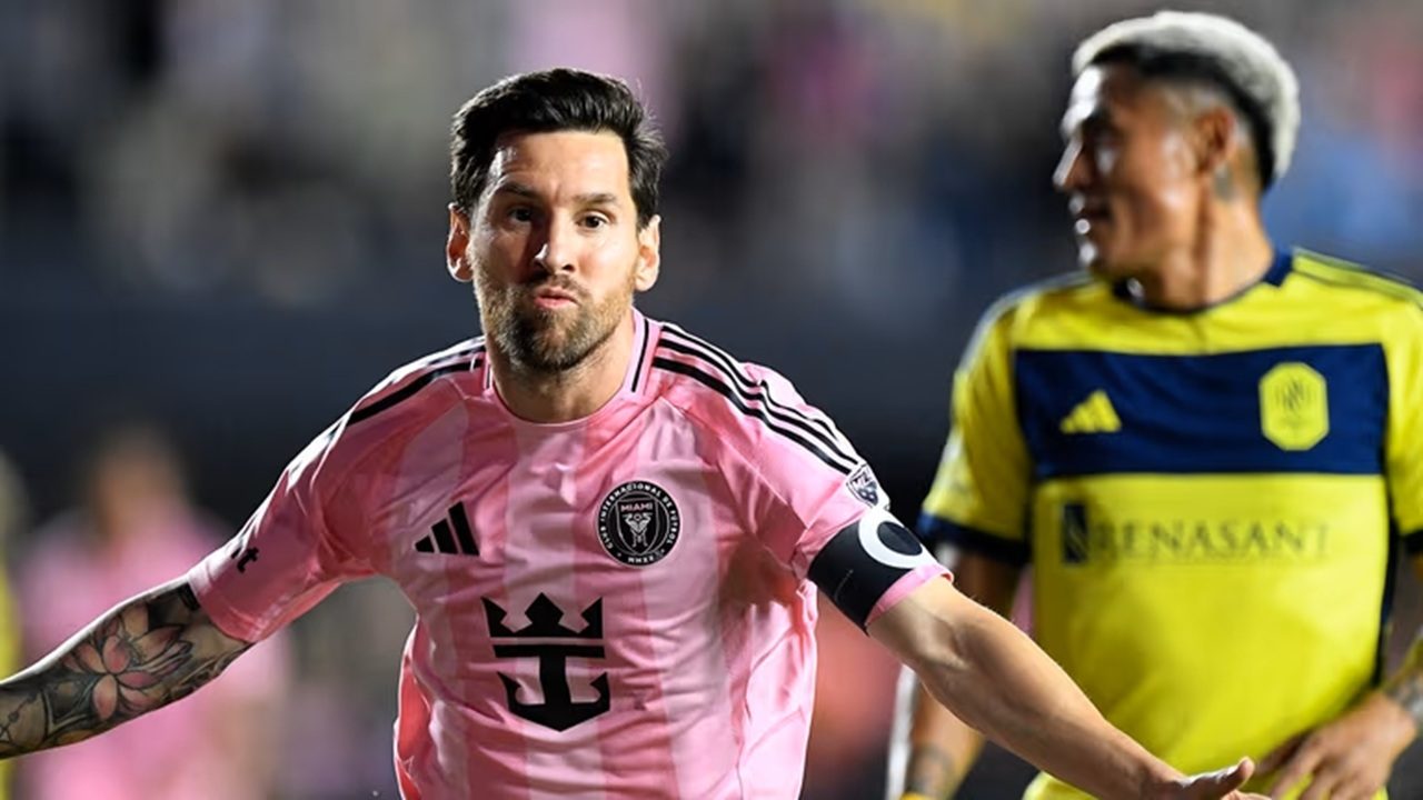 A imagem mostra um jogador de futebol comemorando em campo, vestindo o uniforme rosa do Inter Miami, com o escudo do clube visível no peito e uma braçadeira de capitão no braço esquerdo. Atrás dele, outro jogador com uniforme amarelo e azul observa a cena