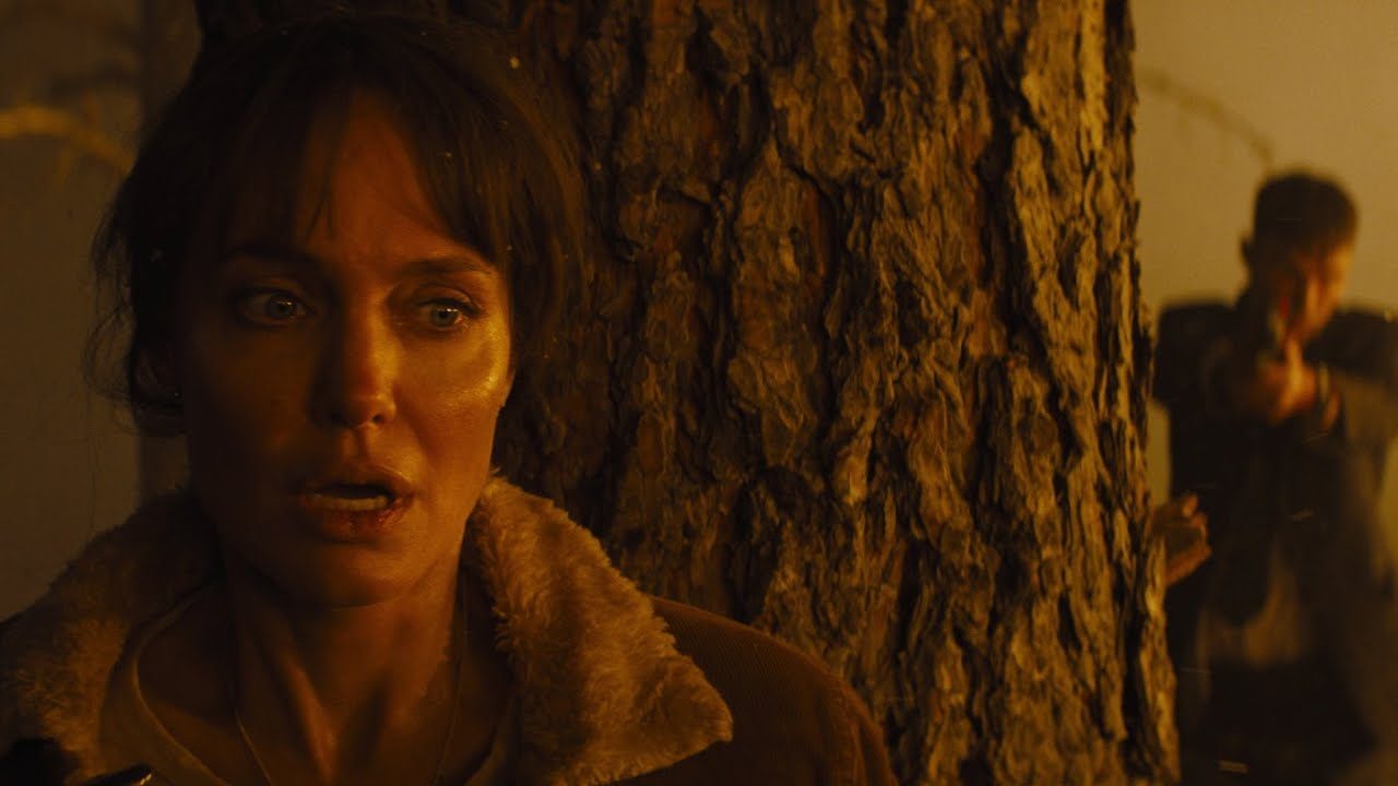 A imagem retrata uma cena tensa do filme "Aqueles Que Me Desejam a Morte" (Those Who Wish Me Dead), estrelado por Angelina Jolie. A personagem interpretada pela atriz aparece encostada em uma árvore, com o rosto sujo de fuligem e uma expressão de medo e tensão, indicando que está em uma situação de extremo perigo.