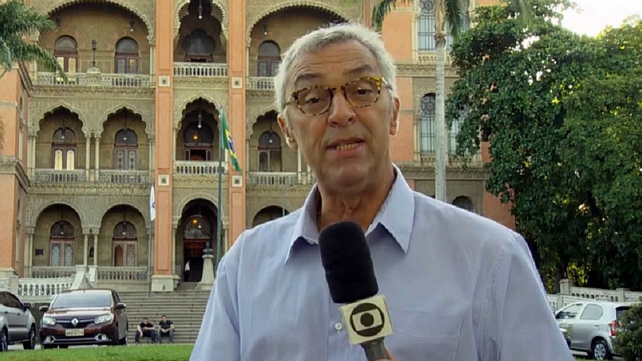 Repórter segura microfone da TV Globo em frente a prédio histórico durante entrada ao vivo na rua