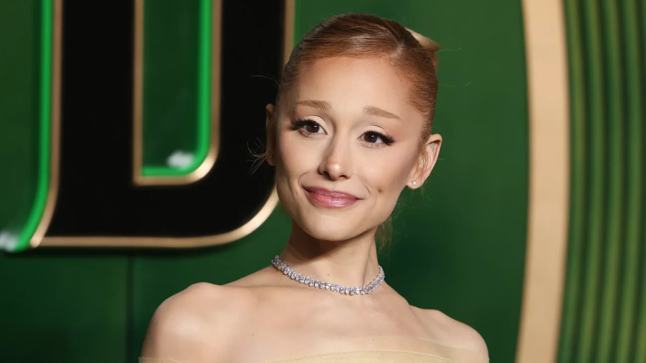 Mulher em evento de tapete vermelho posa diante de um fundo verde com detalhes dourados. Ela usa vestido tomara que caia em tom claro, colar brilhante de pedras e brincos pequenos. O cabelo está preso em um coque elegante, e a maquiagem destaca olhos e lábios com acabamento suave