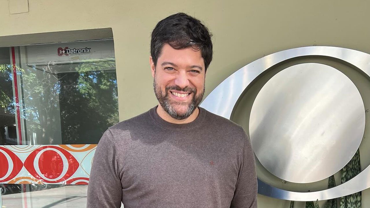 Ariel Jacobowitz sorrindo em frente ao logo metálico da Band na portaria da emissora em São Paulo