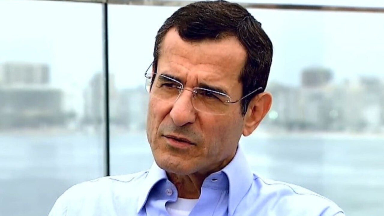 Arnaldo Duran com expressão séria durante entrevista à beira-mar com prédios desfocados ao fundo