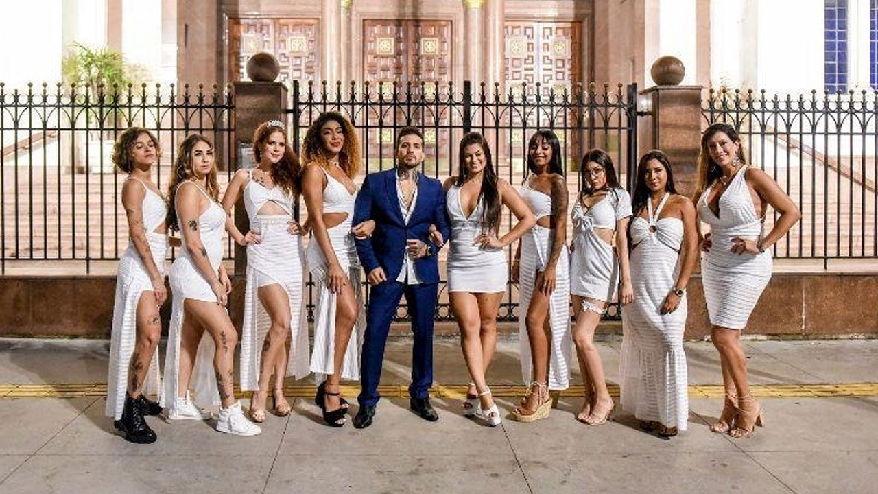 Arthur O Urso posa de terno azul cercado por suas nove mulheres vestidas de branco em frente a uma grade de ferro e um casarão