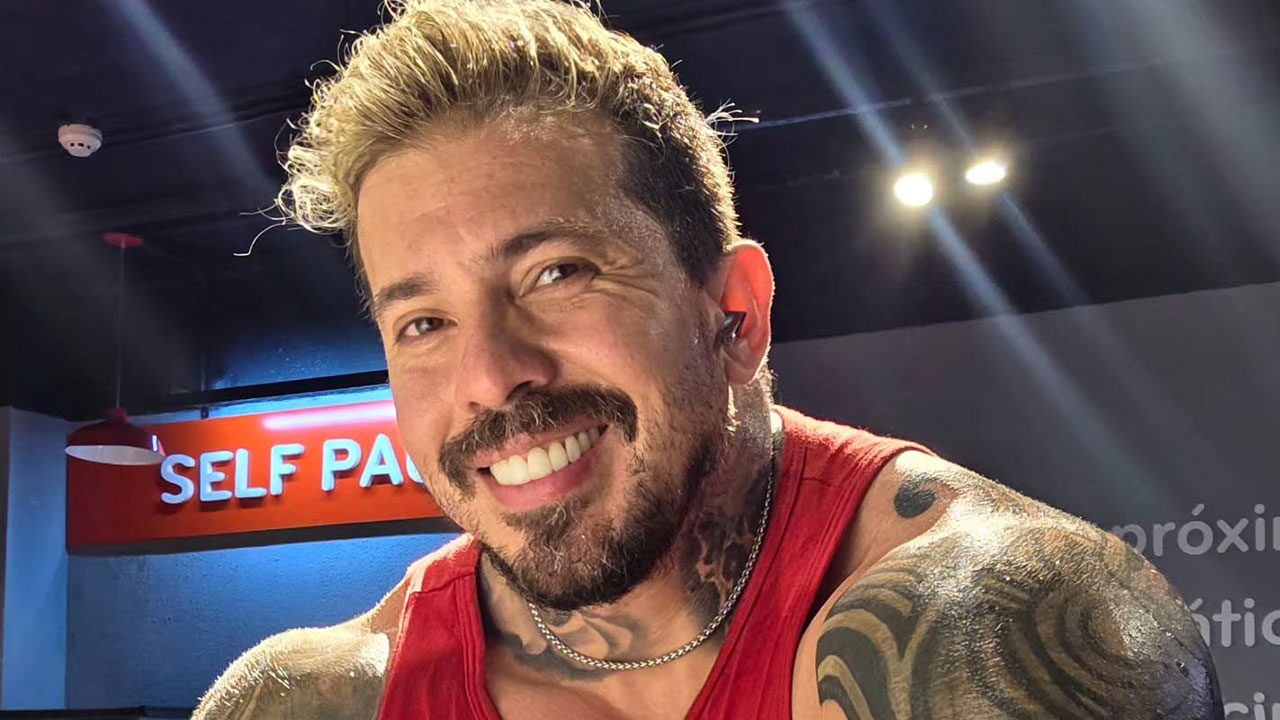 Arthur O Urso sorri em selfie na academia, homem com barba e tatuagens no braço e pescoço, veste regata vermelha e corrente de prata, fundo com luzes