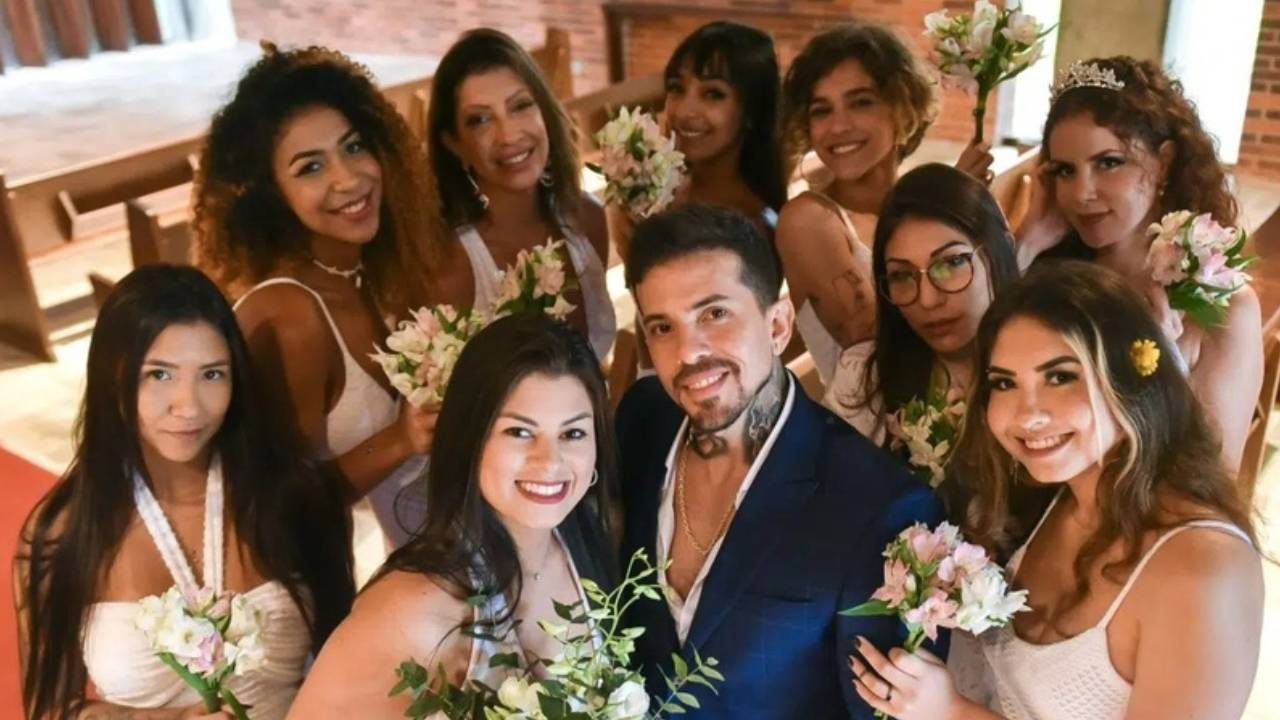 Arthur O Urso posa sorridente de terno azul ao lado das dez mulheres com quem foi casado, todas vestidas de branco e segurando buquês de flores, durante cerimônia simbólica de casamento coletivo