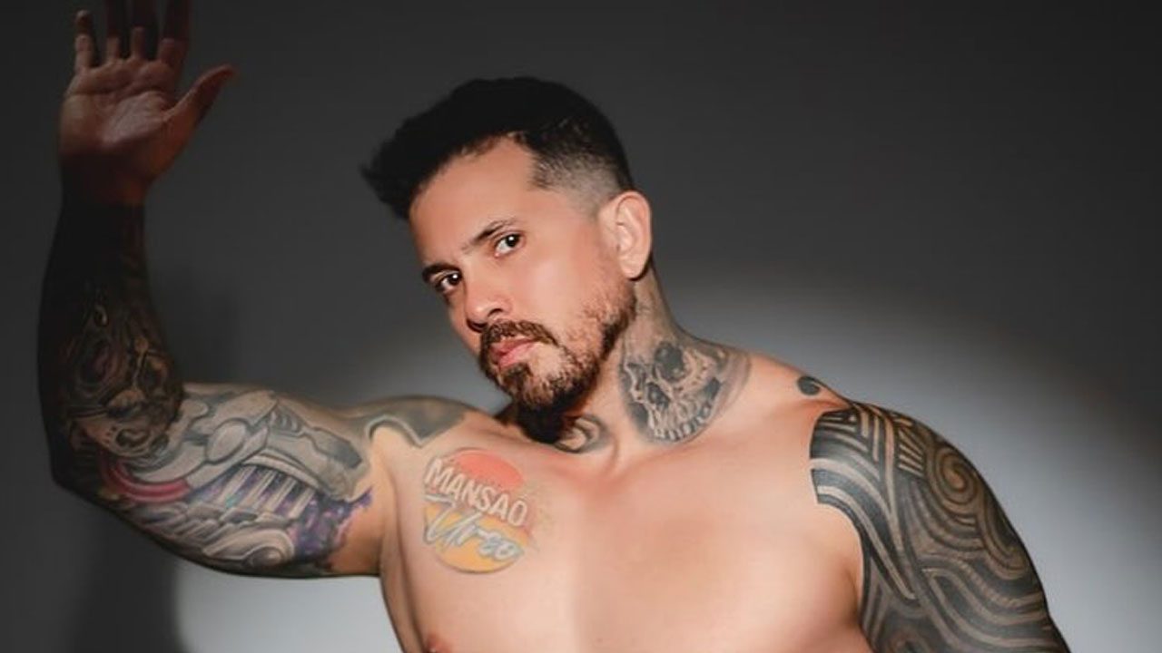 Arthur O Urso sem camisa em ensaio com fundo escuro, exibindo tatuagens nos braços, peito e pescoço