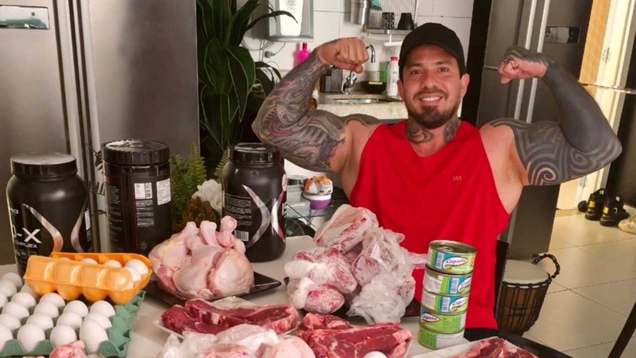 Arthur O Urso posa flexionando os braços em cozinha com mesa cheia de carnes, ovos, suplementos e alimentos ao redor