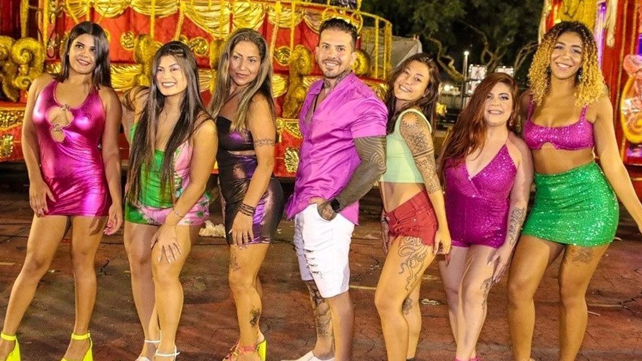 Arthur O Urso sorri de camisa rosa e bermuda branca entre suas seis mulheres com roupas coloridas e brilhantes em um Sambódromo