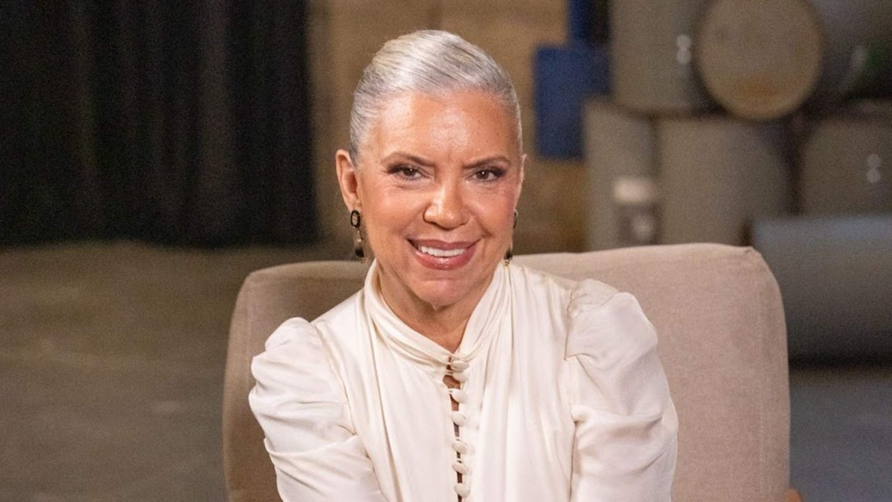 Mulher de cabelos grisalhos presos para trás, usando blusa clara de mangas bufantes, sorri enquanto está sentada em uma poltrona em ambiente interno com fundo desfocado.