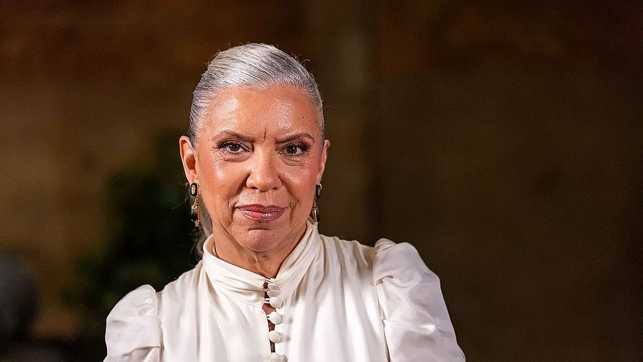 Atriz com cabelos grisalhos presos e blusa branca encara a câmera em cena de novela brasileira