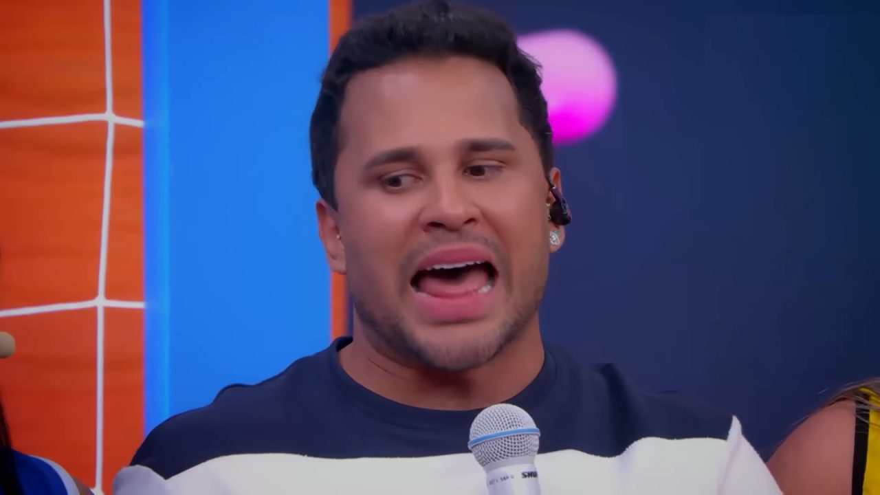 Lucas Guimarães segura um microfone e fala durante gravação do programa Eita Lucas!, usando camiseta azul e branca, fone de ouvido e brinco, com cenário colorido em tons de azul, laranja e rosa ao fundo