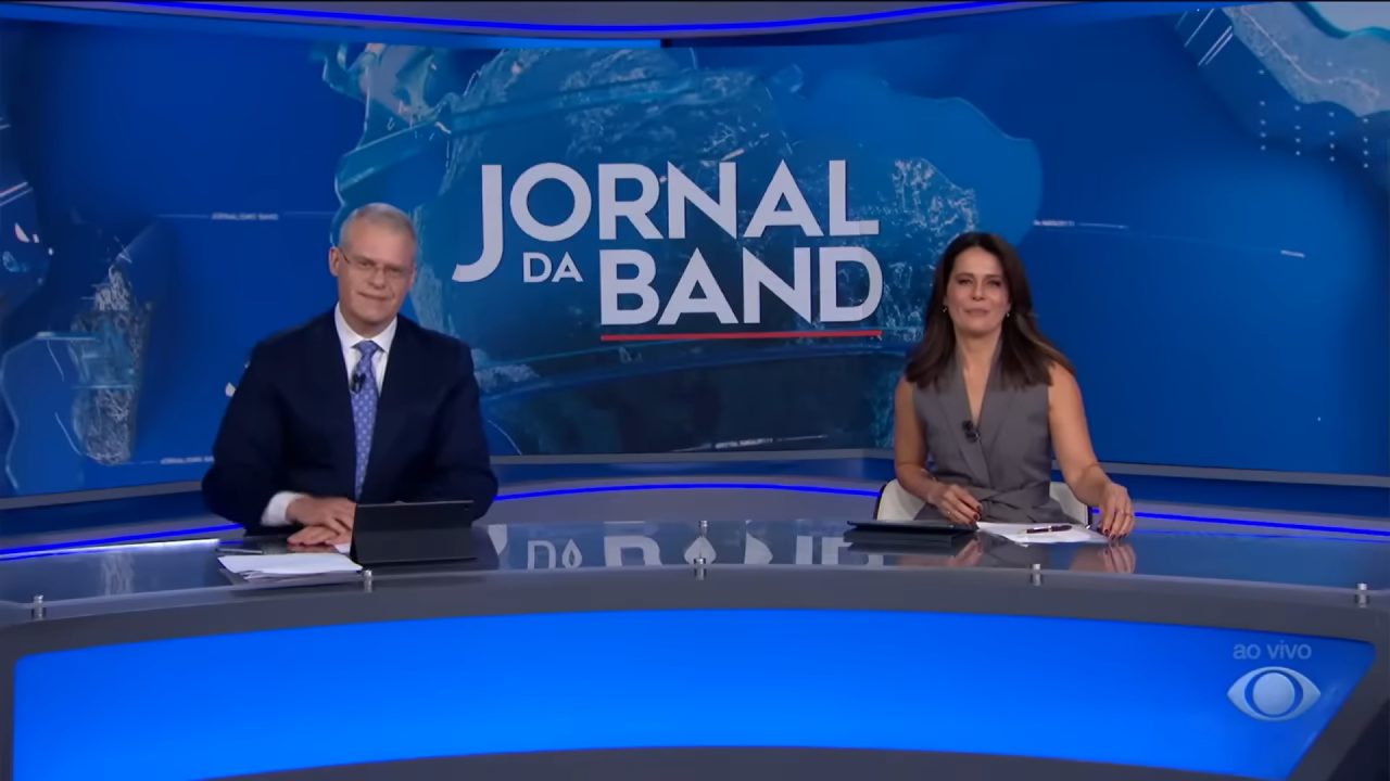 Eduardo Oinegue e Adriana Araújo na bancada do Jornal da Band, em estúdio com telão azul e logotipo do telejornal ao fundo.