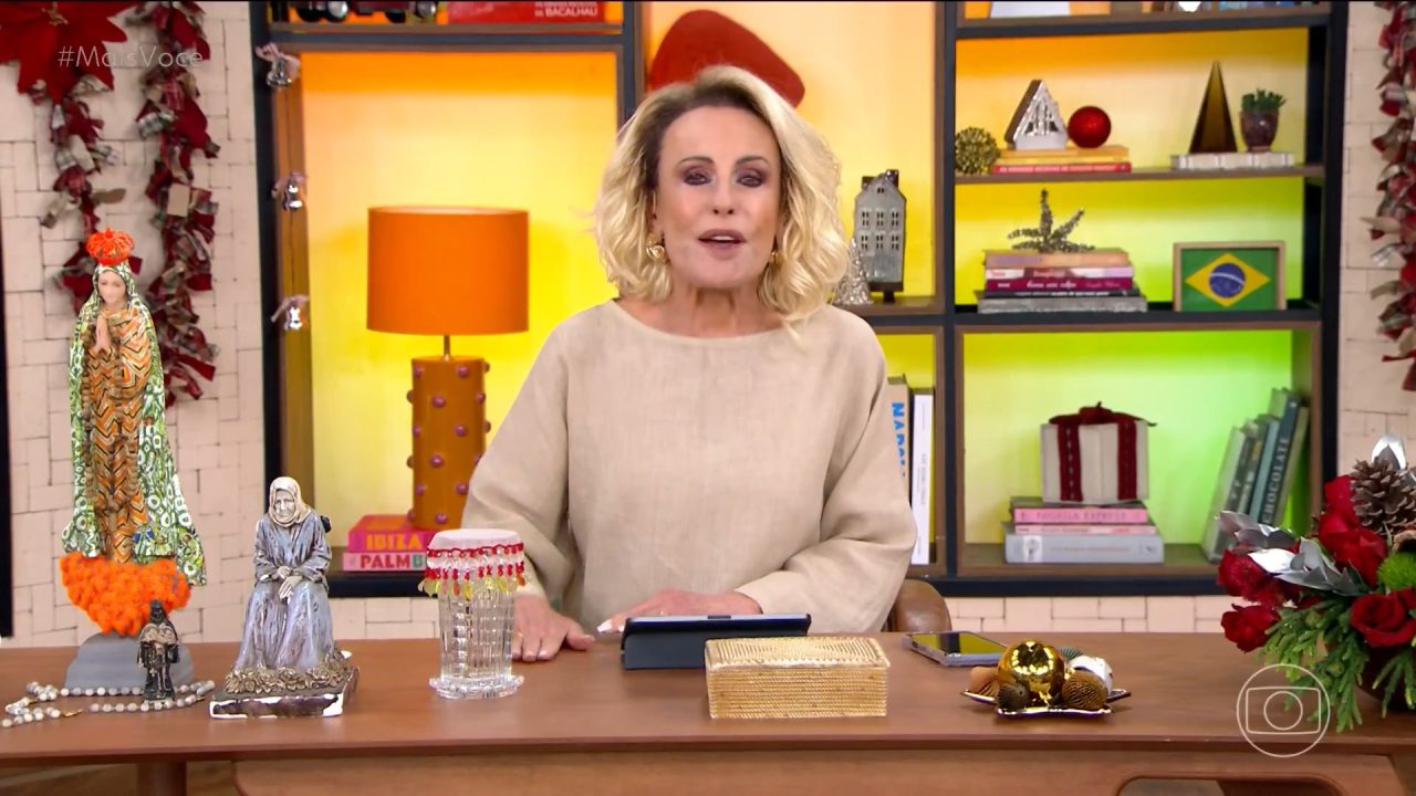 A imagem mostra Ana Maria Braga no estúdio do programa Mais Você, da TV Globo. Ela está atrás de uma mesa decorada com flores, imagens religiosas e enfeites natalinos. Ao fundo, há uma estante colorida com livros, objetos decorativos e uma bandeira do Brasil. No canto superior esquerdo, aparece a hashtag #MaisVocê, identificando o programa matinal