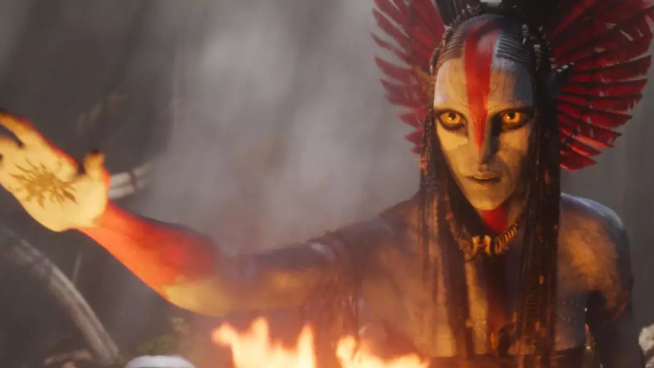 A imagem mostra uma personagem azul com olhos dourados e expressão intensa, com o corpo pintado em tons de vermelho e branco. Ela estende o braço em direção a algo fora de quadro, com um símbolo dourado desenhado na mão. Usa um adorno de cabeça com penas vermelhas e pretas e está cercada por chamas, sugerindo uma cena ritualística ou mística em meio a um ambiente de floresta.