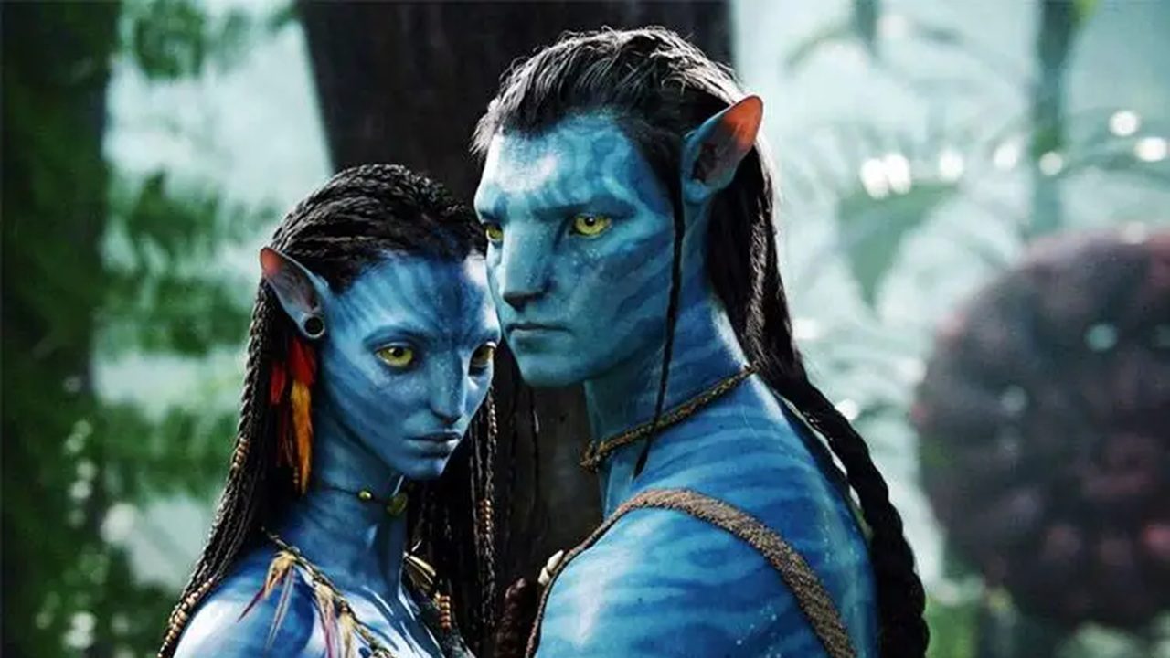 A imagem mostra dois personagens azuis de aparência alienígena com olhos amarelos e orelhas pontudas, em um cenário de floresta exuberante. Eles pertencem à espécie Na’vi do filme “Avatar”, dirigido por James Cameron.