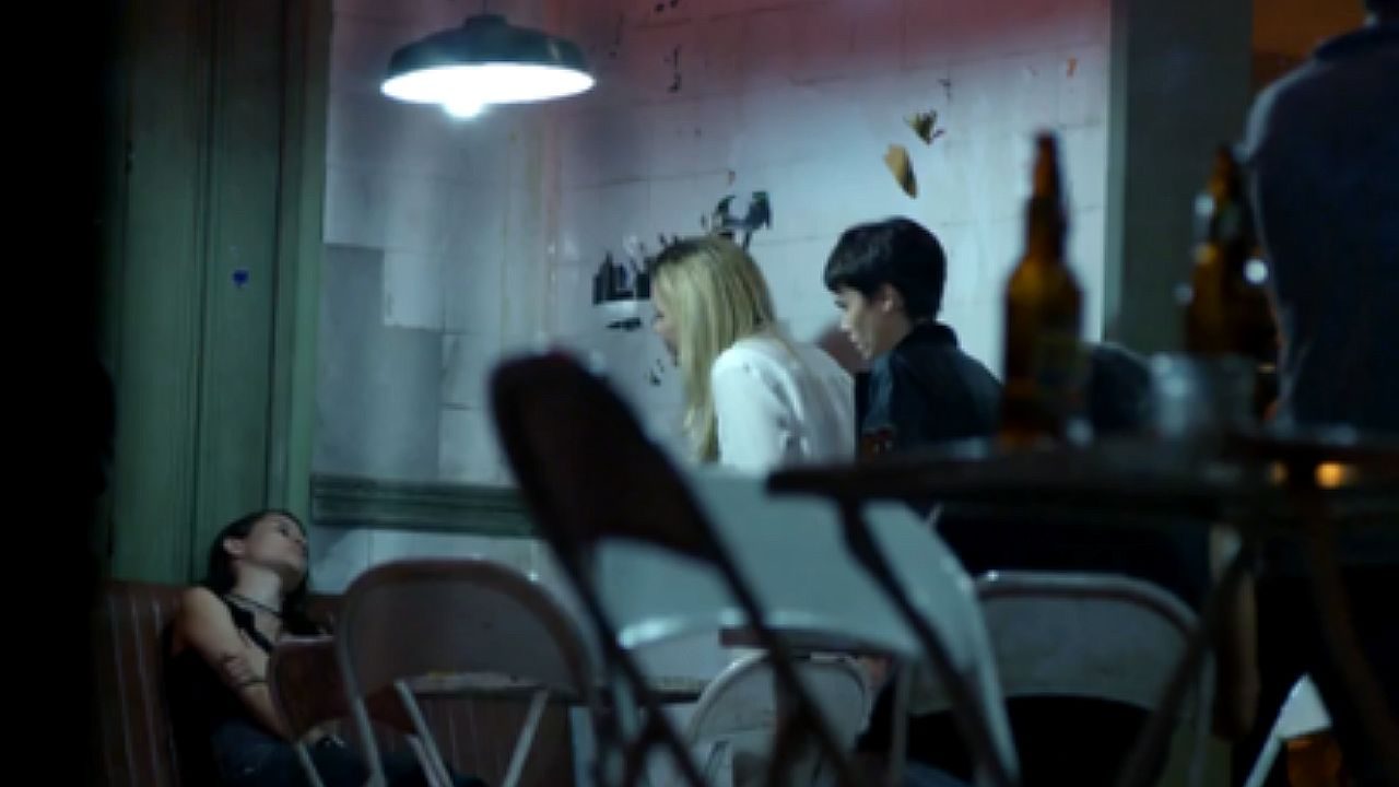 Mulher desacordada ou ferida permanece caída em cadeira enquanto mulher loira e menino observam a cena em ambiente fechado com iluminação baixa e mesas ao redor