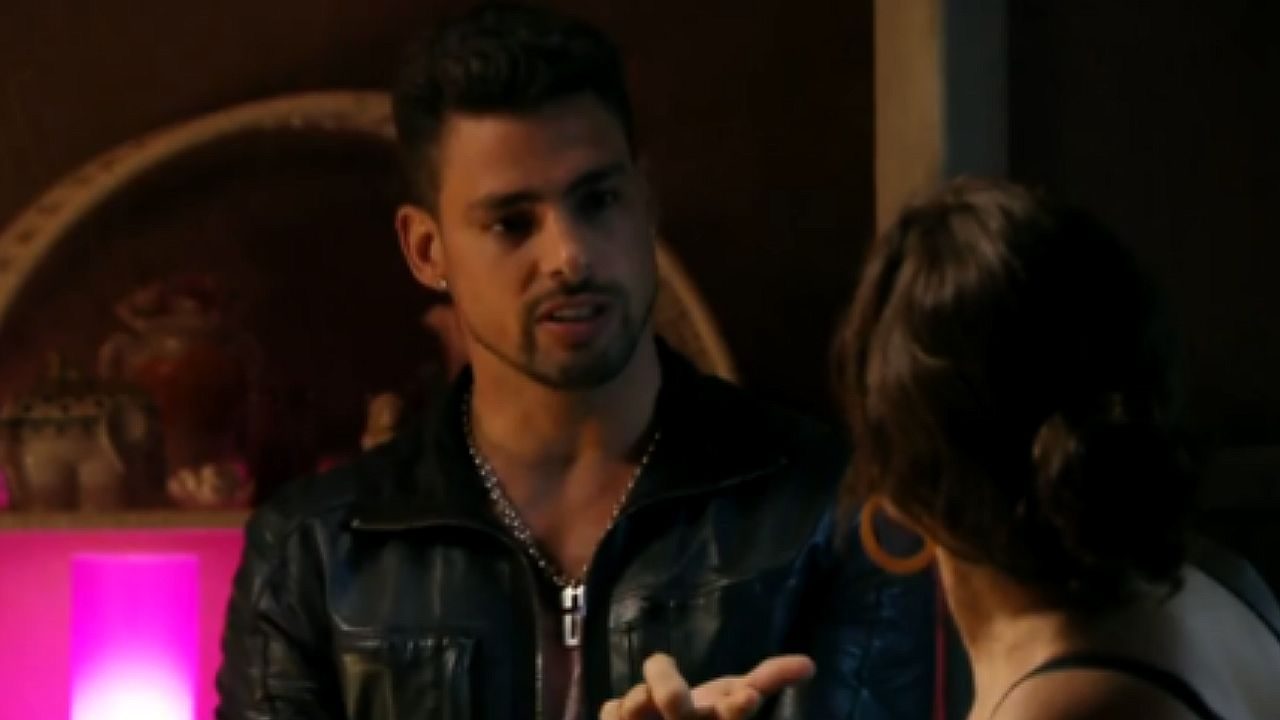 Homem de jaqueta de couro conversa com mulher em ambiente iluminado por luzes coloridas em cena de novela jovem