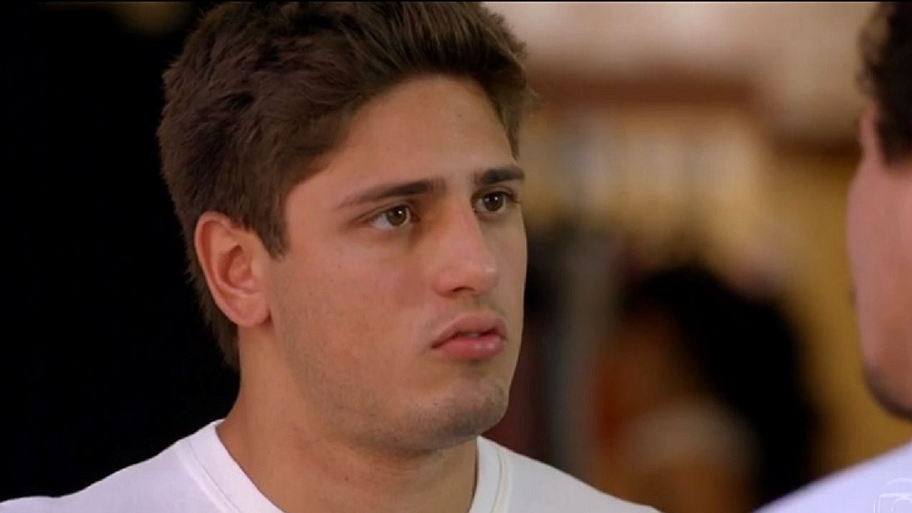 Jovem com expressão tensa encara outra pessoa em cena de novela ambientada em interior com fundo desfocado