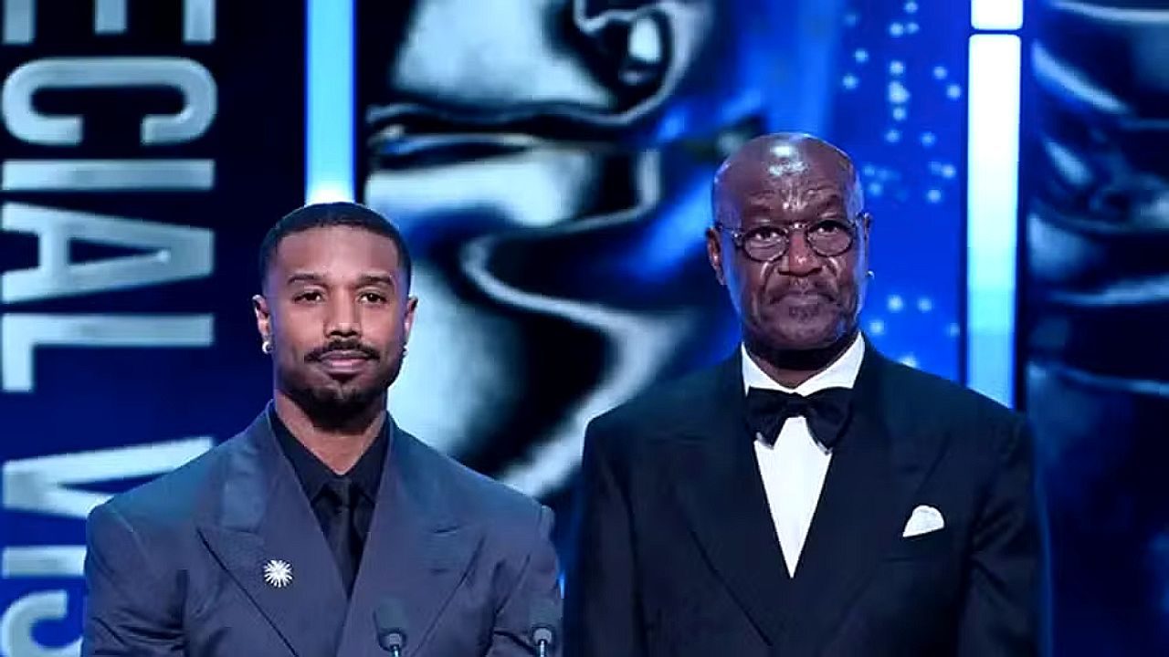 Dois homens vestindo ternos formais apresentam-se em palco durante premiação. À esquerda, um homem de terno azul-escuro fala ao microfone; à direita, outro homem usa smoking com gravata borboleta. Ao fundo, iluminação azul e a imagem ampliada de uma estatueta indicam cerimônia de gala.