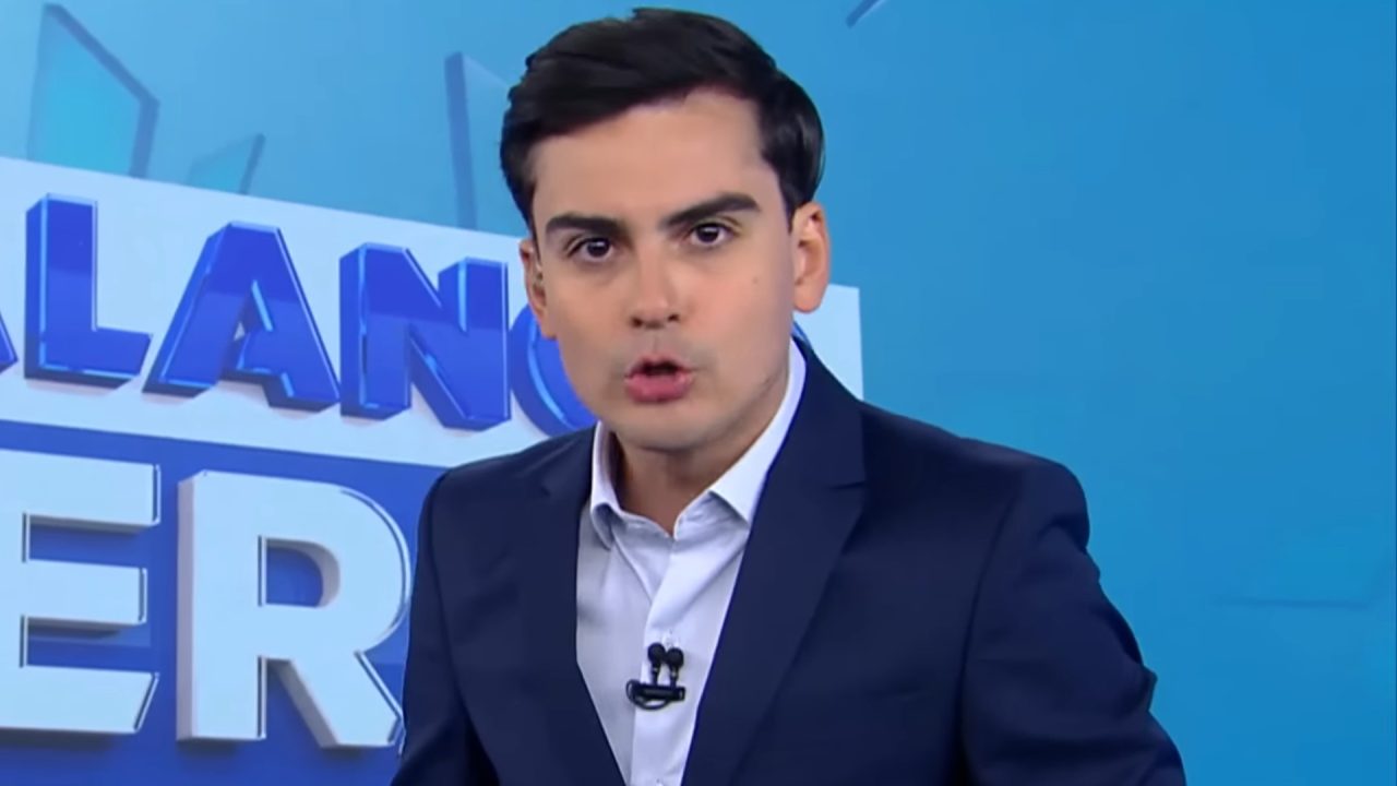 Dudu Camargo, de terno escuro e camisa clara, aparece no estúdio do Balanço Geral com fundo azul, falando de forma séria para a câmera. Ao fundo, há letras grandes do nome do programa parcialmente visíveis.