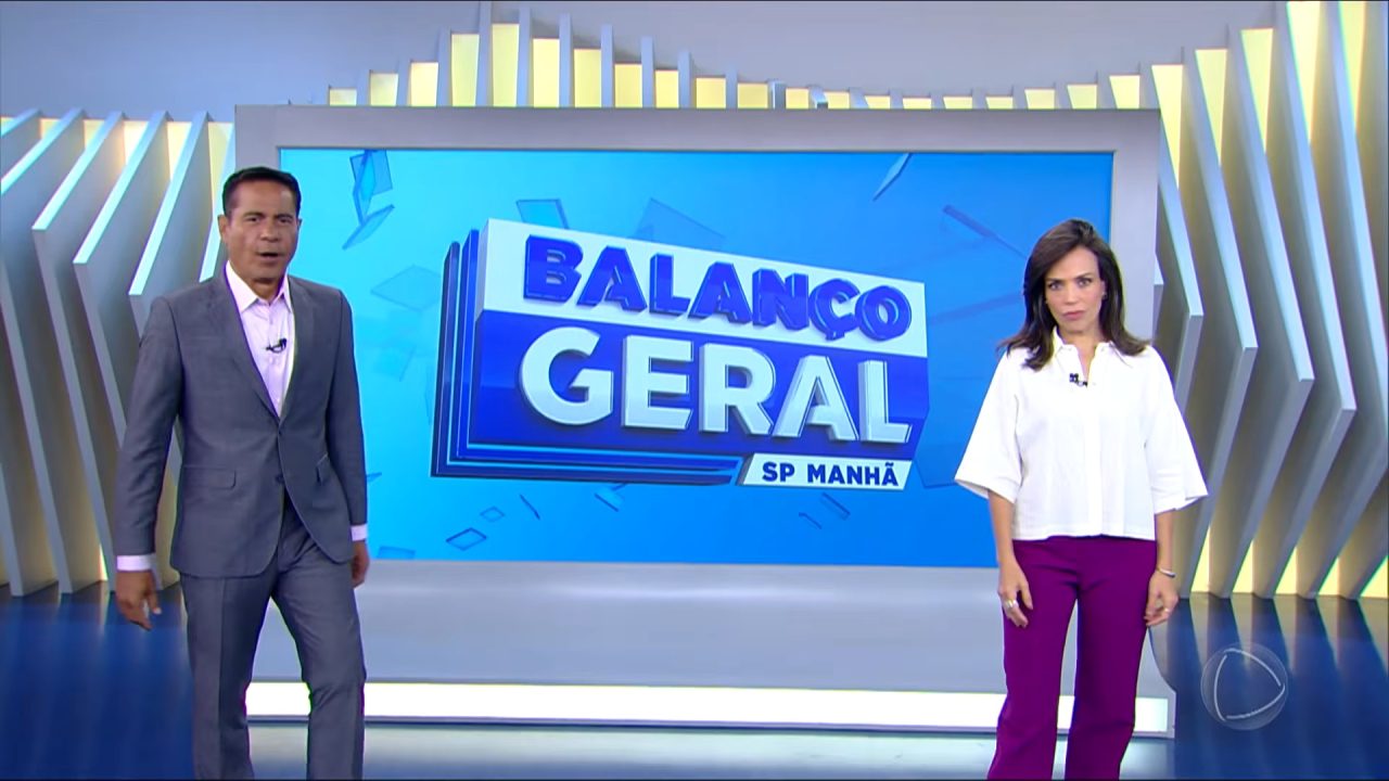 Willian Leite e Fabiana Oliveira, apresentadores do Balanço Geral Manhã, estão em pé no estúdio diante de telão com logotipo do programa.